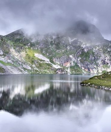 Scenic view of a tranquil lake surrounded by misty mountains in the Rila Mountains, Bulgaria.