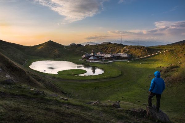 Back view of anonymous male traveler in warm clothes standing on stone and admiring picturesque view of holy alpine Prashar lake and temple located in India