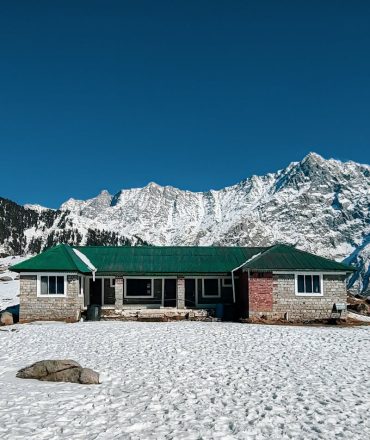 Snow-covered lodge with green roof against majestic mountain backdrop.